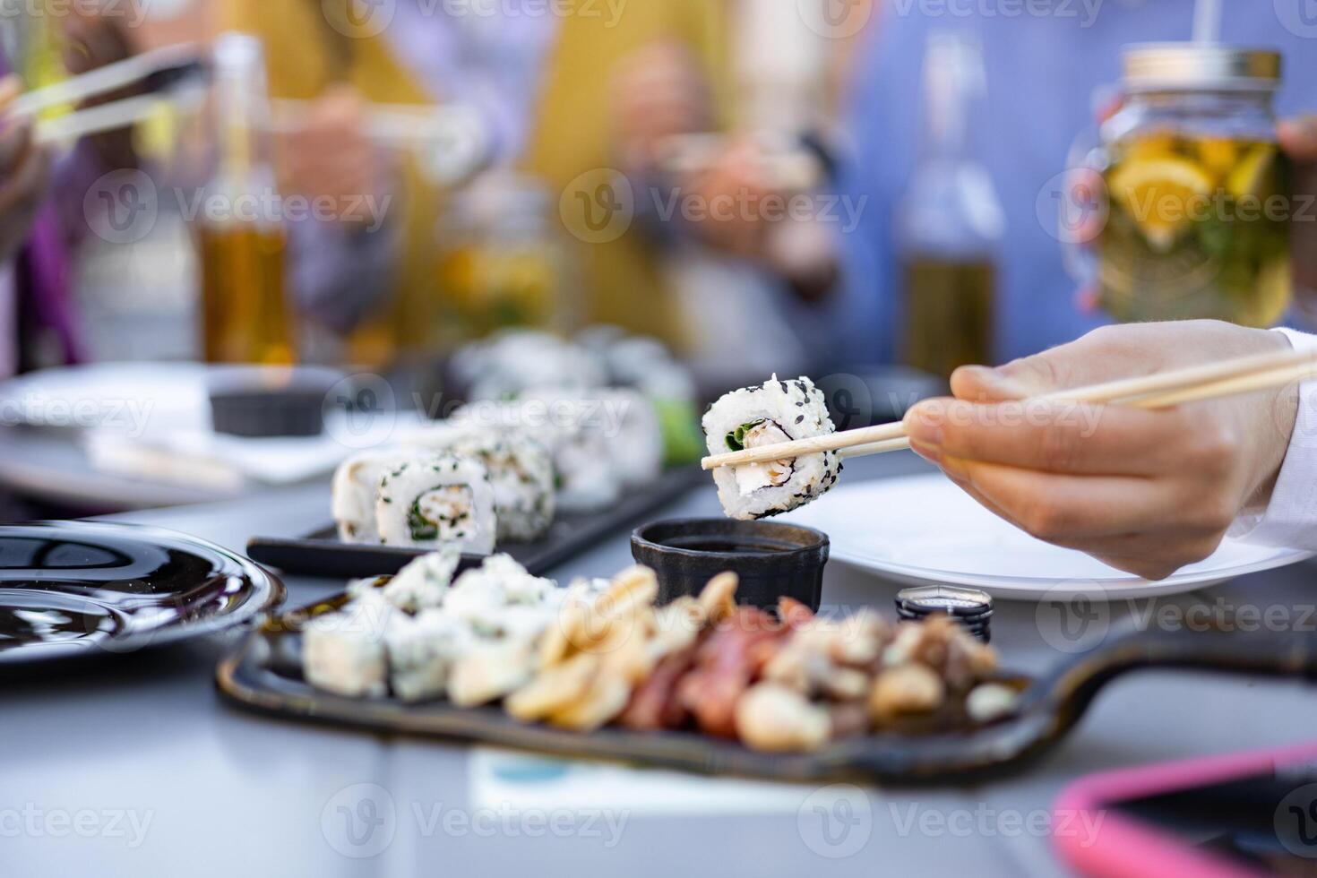Close-up of a hand using chopsticks to eat sushi from a table set with other food and drinks. Friends enjoy the meal. photo
