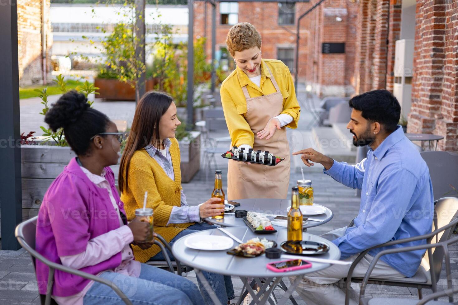 A server presents sushi to a group of friends sitting at an outdoor restaurant table, drinks and food are visible. photo