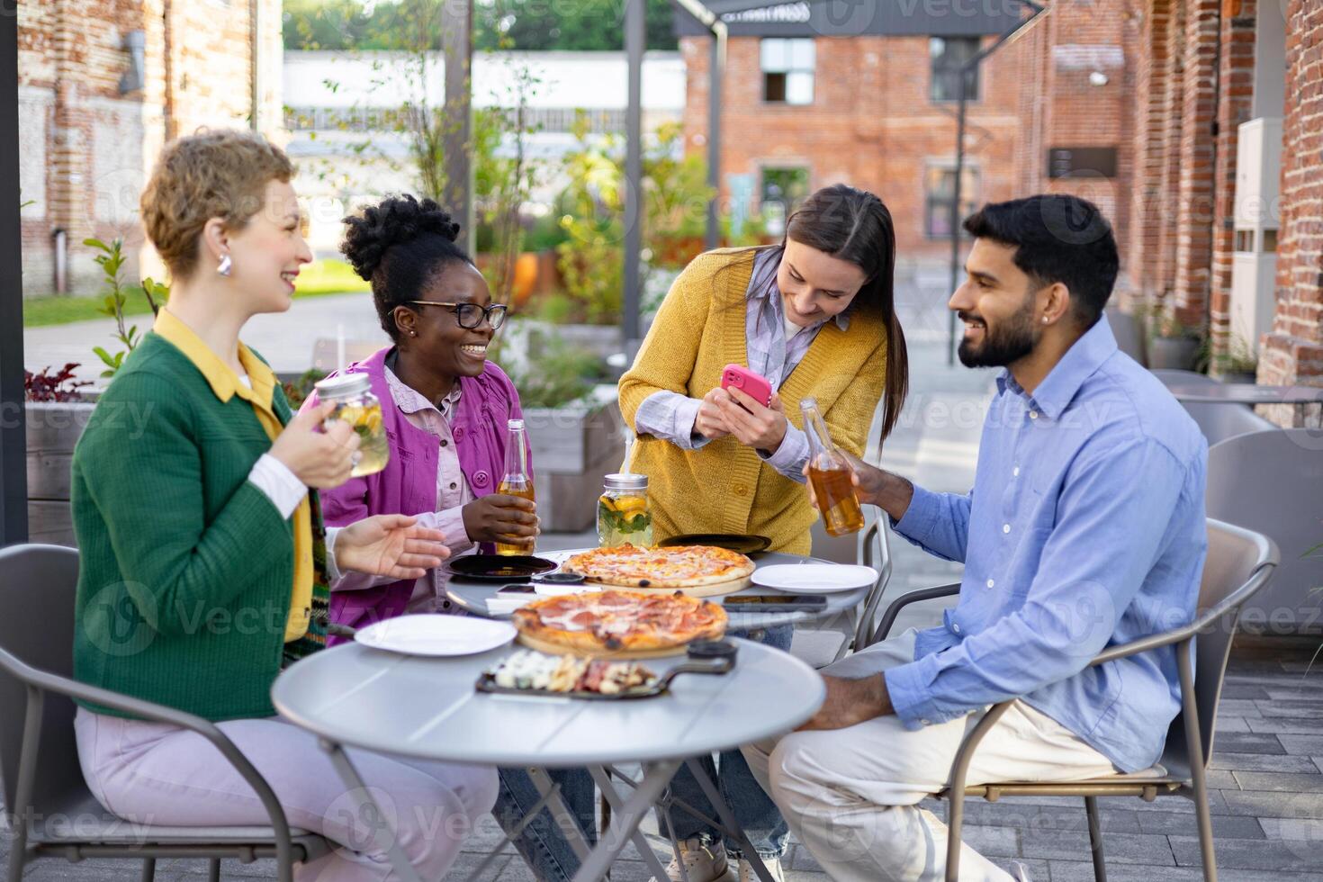 A diverse group of friends enjoys pizza and drinks at an outdoor restaurant, capturing a moment together with a photo. photo