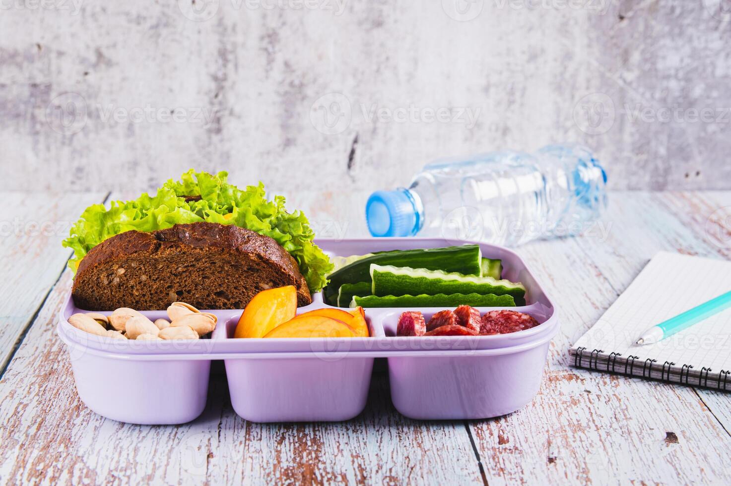 Lunch box with food, bottle of water, notebook and pen on the table photo