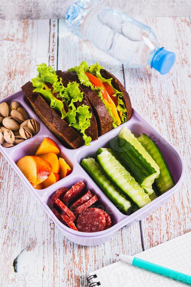 Lunchbox with sandwich, chorizo, cucumber and peach, bottle of water on table vertical view photo
