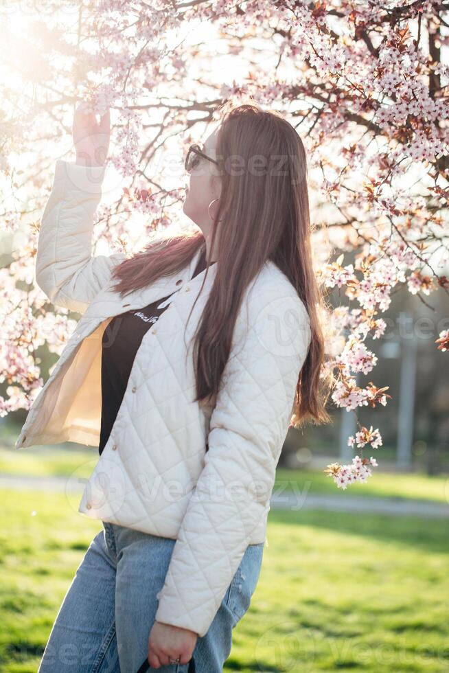 Woman enjoying springtime blossoms in a park while wearing a stylish jacket and sunglasses on a sunny day photo