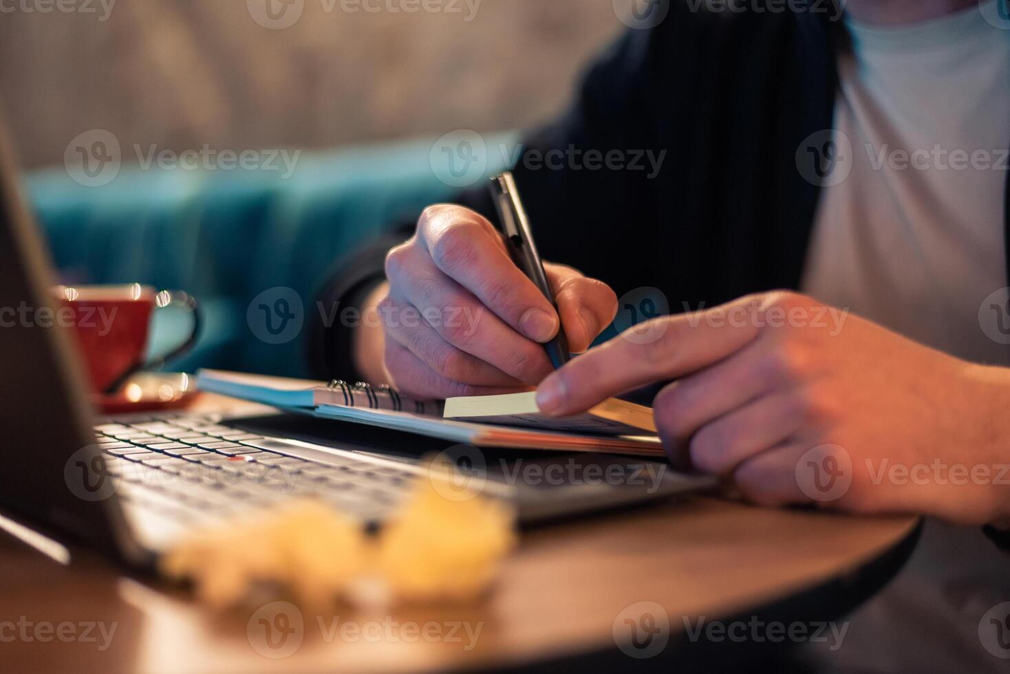 Close cropped image of a business man writing in a notebook with crumpled papers on the desk while working on a laptop or a male student studying in a modern interior. Concept of a online learning. photo