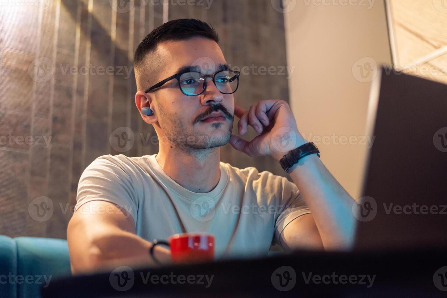 Focused young man wearing glasses and wireless earbuds, watching a webinar, having a call with a client or a business conference meeting on a laptop in a modern interior with ambient light. photo