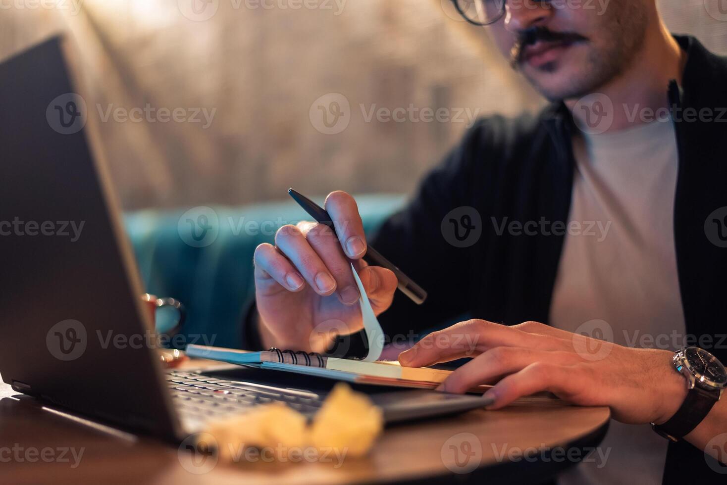 Close up of a man working late on a laptop, writing on a sticky note and pen, with crumpled papers on the desk, concept of planning, brainstorming, or creative work. photo