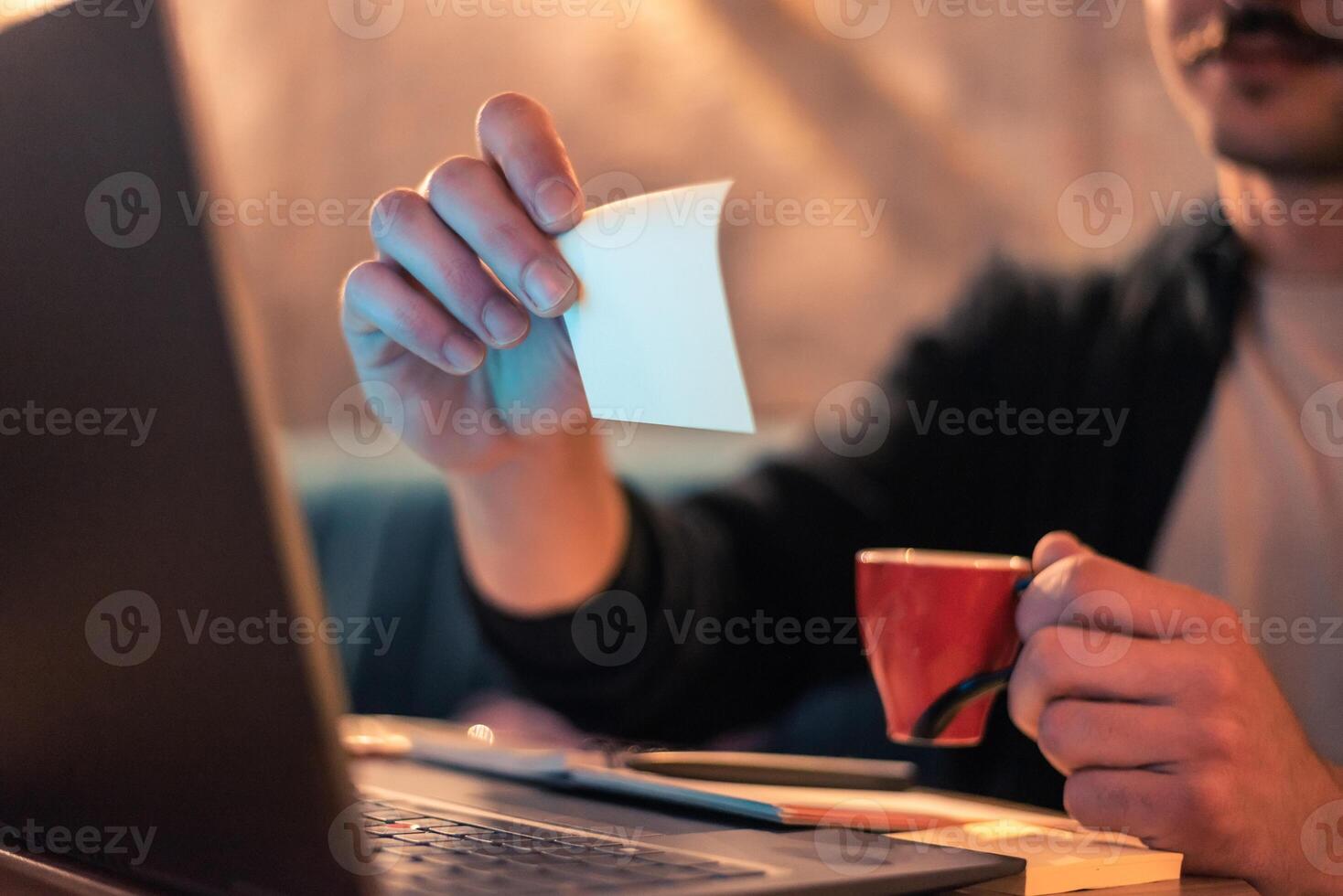Close up of a man working late on a laptop in a modern interior with ambient light, holding a sticky note and a cup of coffee, concept of planning, brainstorming, or creative work. photo