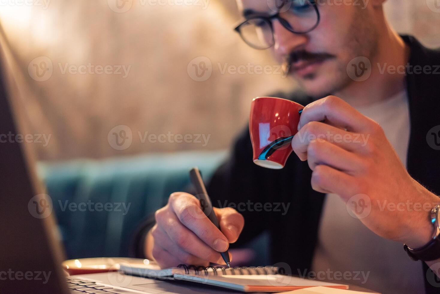 Close cropped image of a young man writing in a notebook and holding a cup of coffee while working late on a laptop or a male student studying in a modern interior with evening interior light. photo