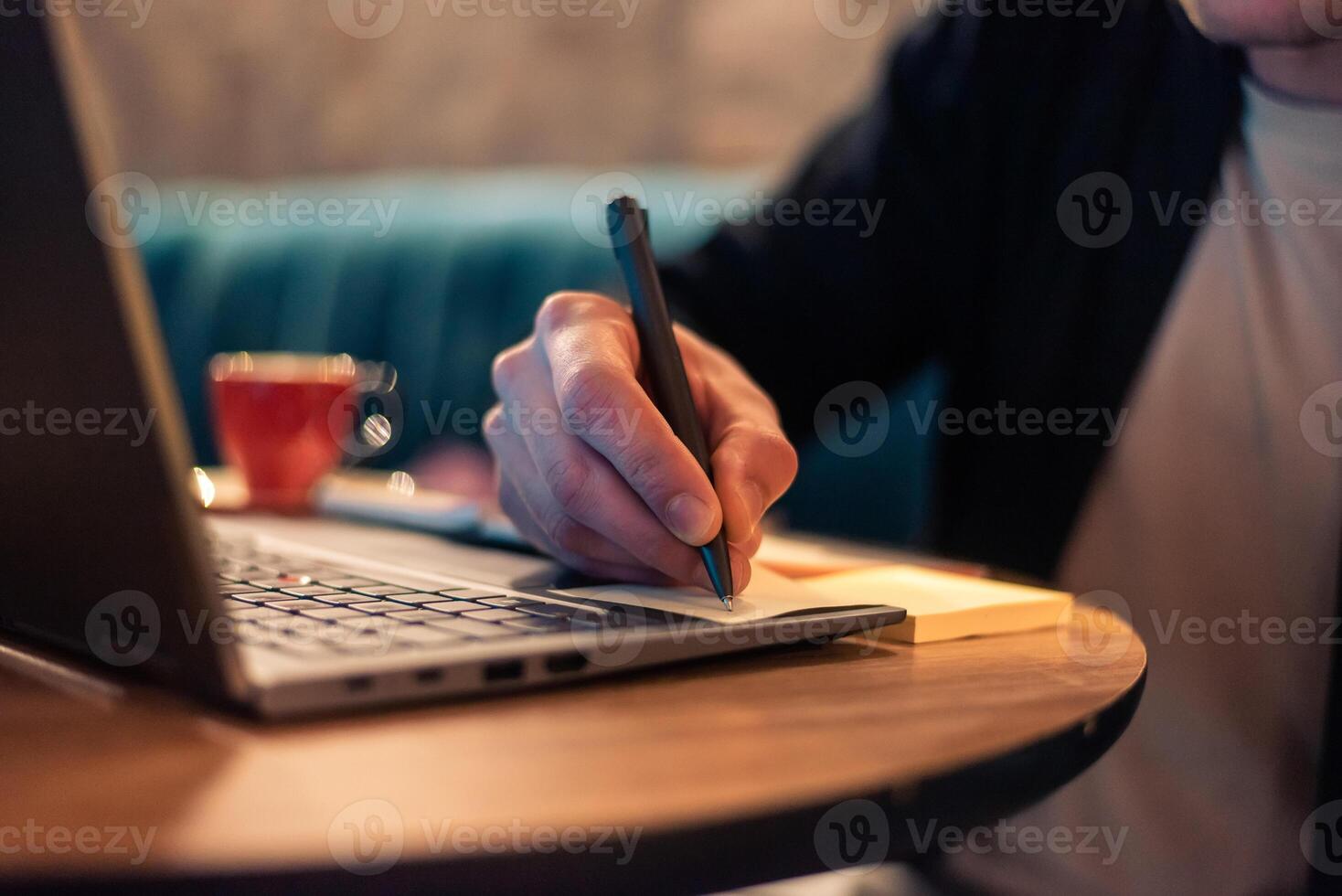 Close cropped image of a business man writing in a sticky note while working on a laptop or a male student studying in a modern interior. Concept of a business planning, strategy and online learning. photo