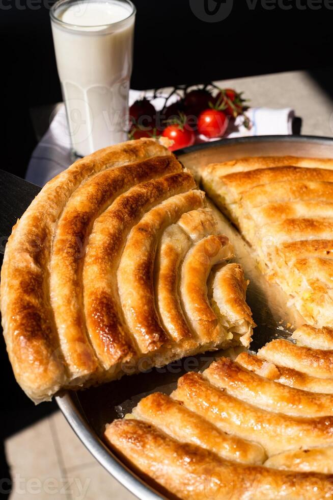 Close-up of a freshly baked spiral pie with golden crust, served in a round tray. A slice is being lifted, with a glass of yogurt and fresh cherry tomatoes in the background, evoking a homemade meal. photo