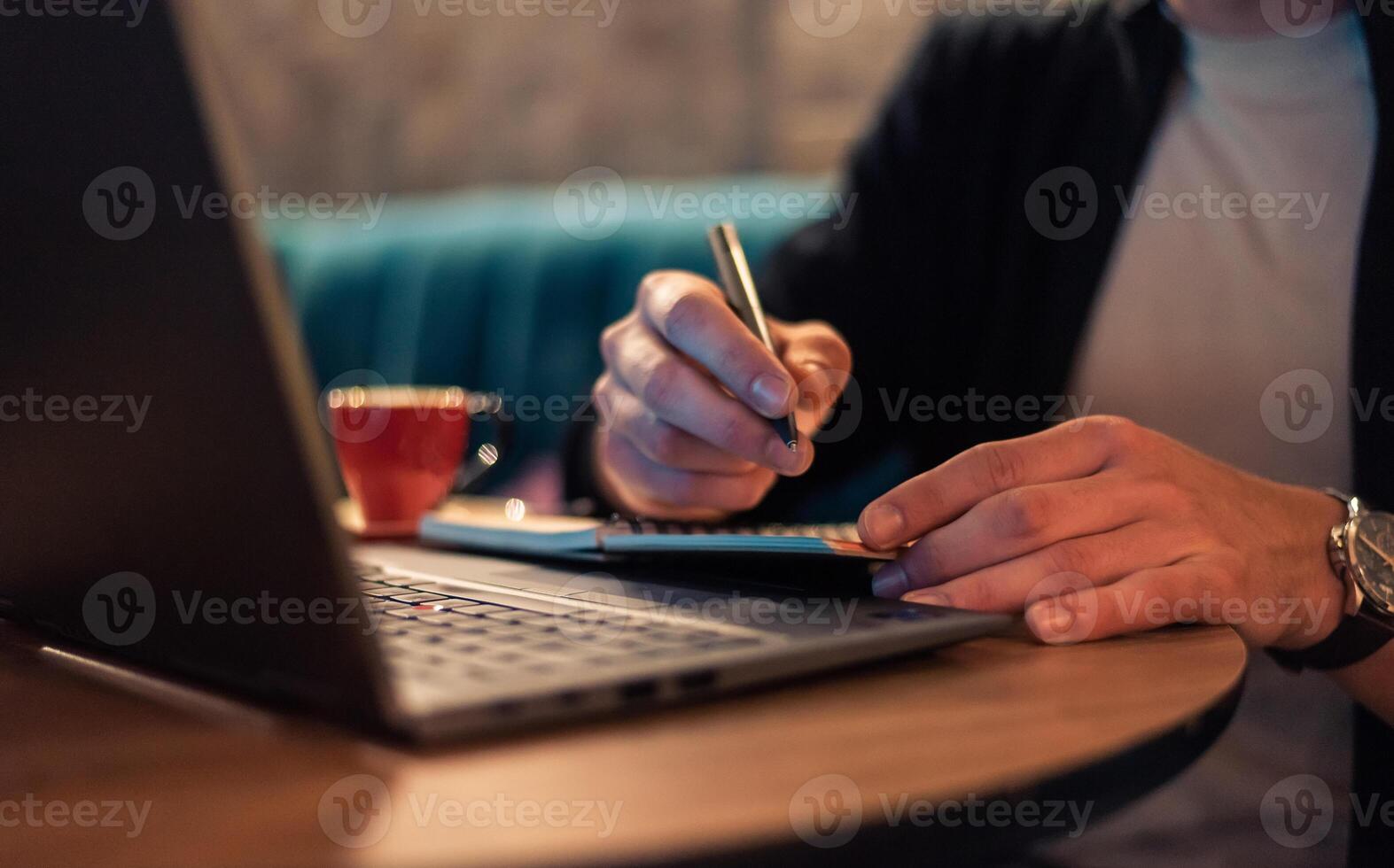 Close cropped image of a businessman writing in a notebook while working on a laptop or a male student studying in a modern interior. Concept of a business planning, strategy and online learning. photo
