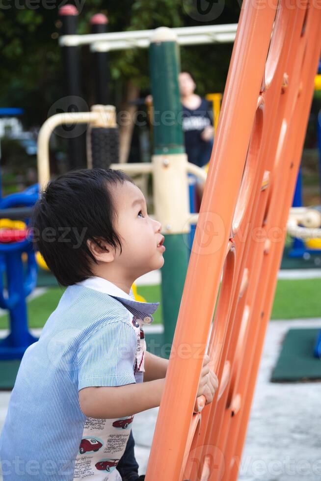 Little boy playing happily on the playground in the summer sun. Asian kid climb the orange play equipment for climbing, play and development of small and large muscles, decision making and courage. photo