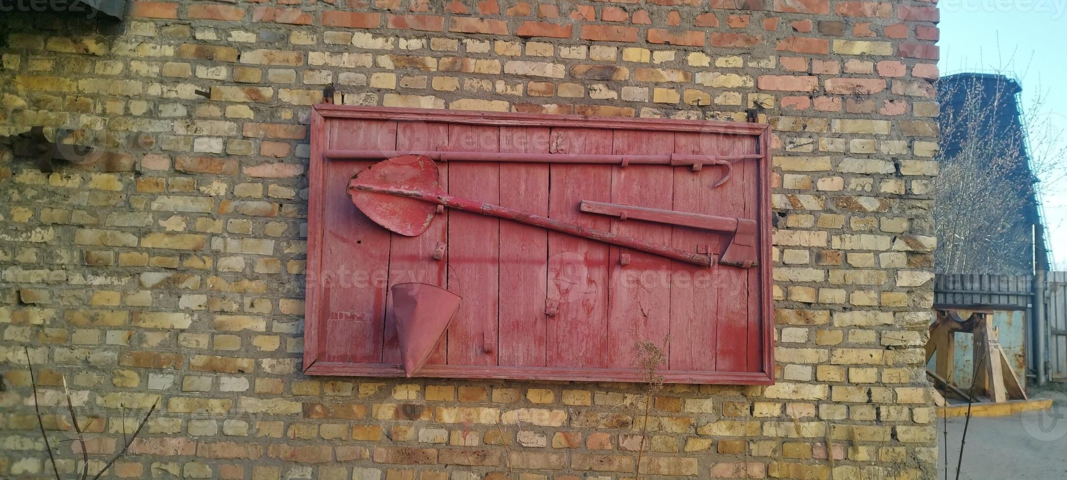 Vintage fire fighting tools displayed on a red wooden board, mounted on a brick wall. Axe, shovel, bucket, and hook. Fire shield. Displayed outdoors against a brick wall. Red wooden fire shield. photo