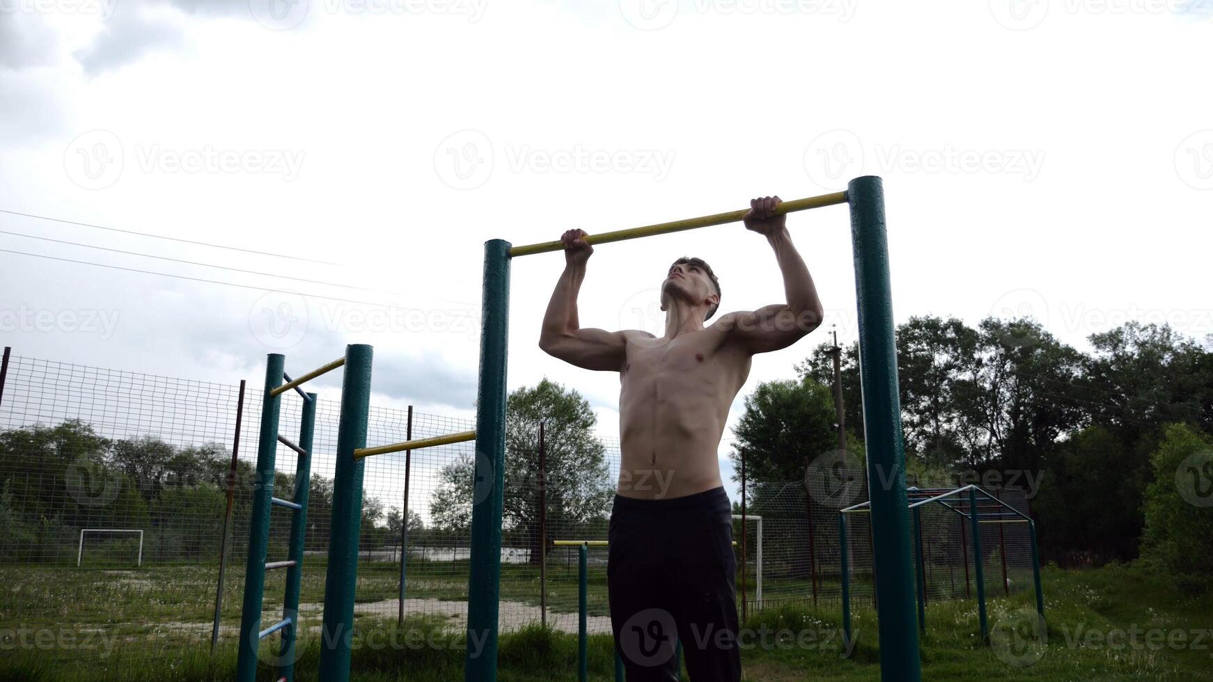 Muscular athlete doin pull ups on horizontal bar at sports ground. Young shirtless sportsman exercising at nature. Strong sporty man training outdoor. Concept of healthy and active lifestyle. Slow mo photo
