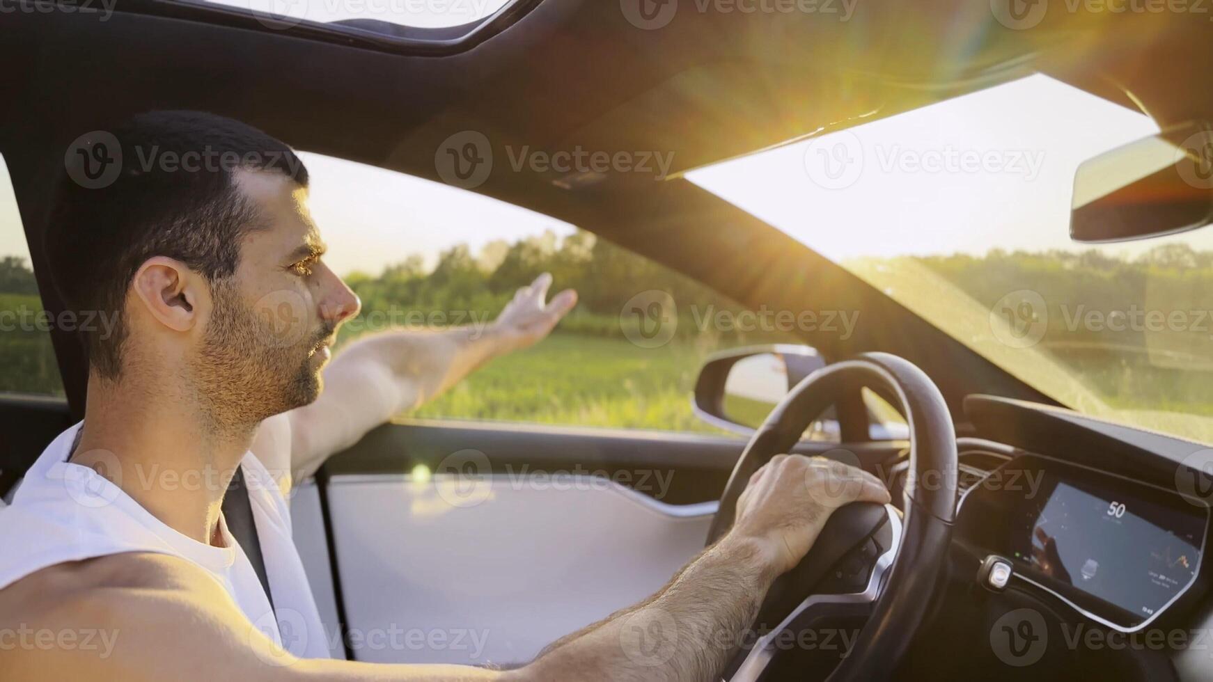 Profile young handsome man driving a modern electric car with sunlight at background. Young male driver riding on electrical vehicle at countryside. Concept of journey and trip. Close up photo