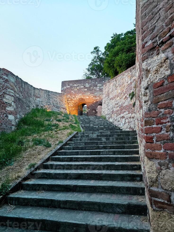 Stone steps lead to an illuminated archway in an old fortress. The path is flanked by historic brick and stone walls, leading the viewer into a serene twilight setting photo