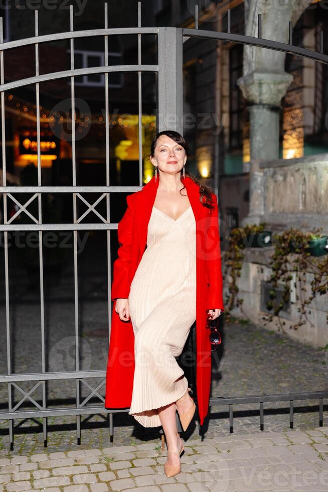 Woman wearing red coat and beige dress standing by a gate at night photo