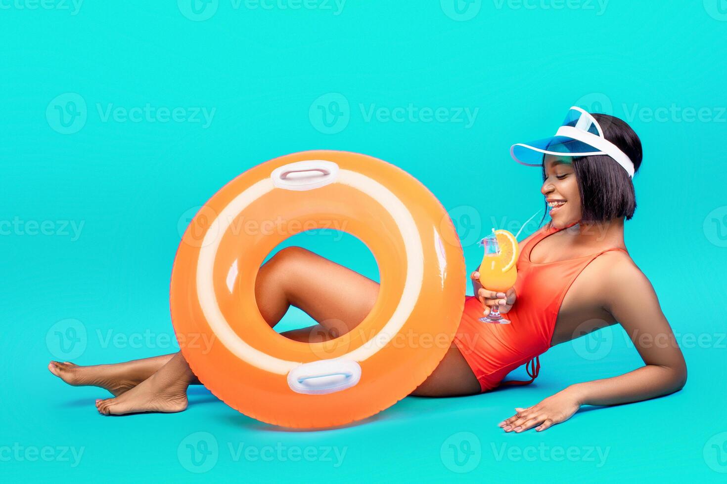 Pool party time. Millennial black woman with fresh tropical cocktail and inflatable ring having fun summer vacation, lying on blue studio background, full length shot photo