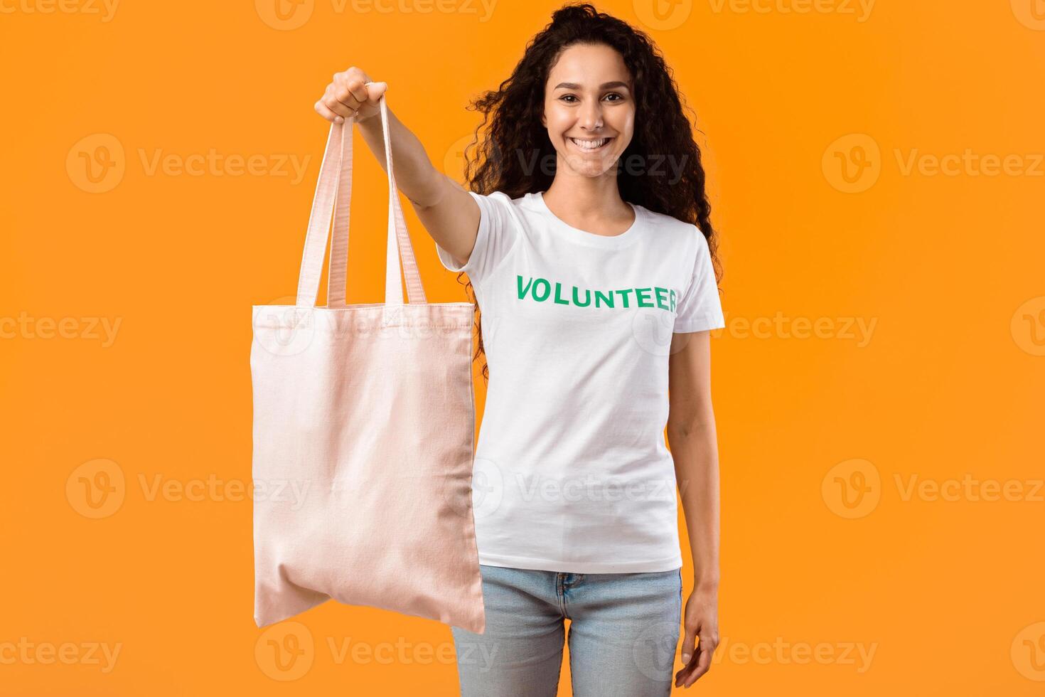 Smiling Volunteer Lady Showing White Eco Bag Standing In Studio Over Yellow Background. Ecologigal Activist Offering Textile Tote Handbag For Shopping. Don't Use Plastic Bags. Fashion And Ecology photo