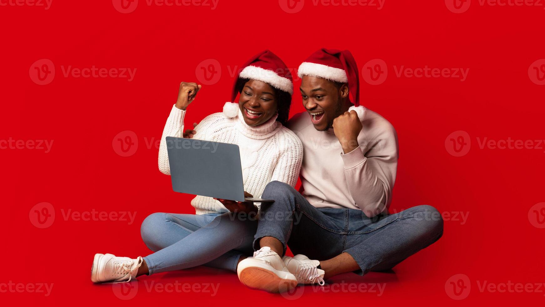Christmas Offer. Excited black couple in santa hats celebrating success with laptop over orange background, cheerful african spouses sitting with computer and raising fists with joy, copy space photo