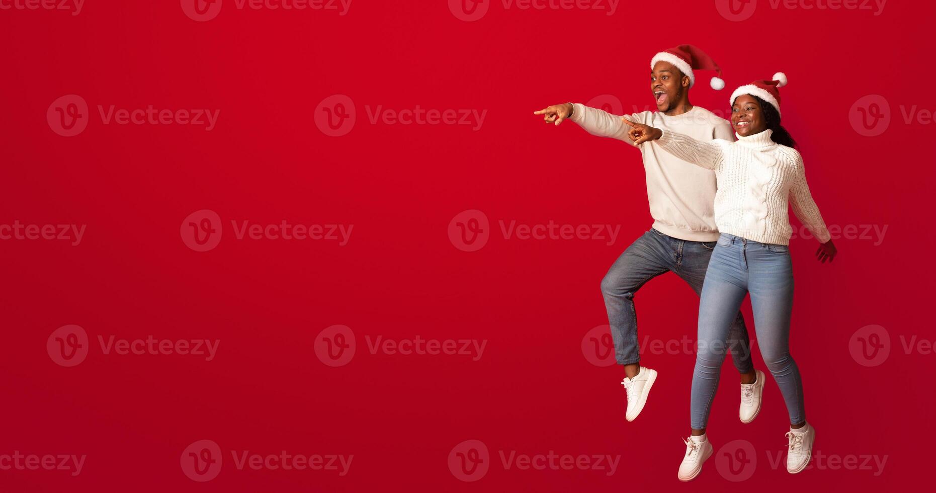 Promo Offer. Cheerful Black Couple In Santa Hats Pointing At Copy Space, Demonstrating Free Place On Orange Background For Your Christmas Holiday Advertisement, Jumping Together And Having Fun photo