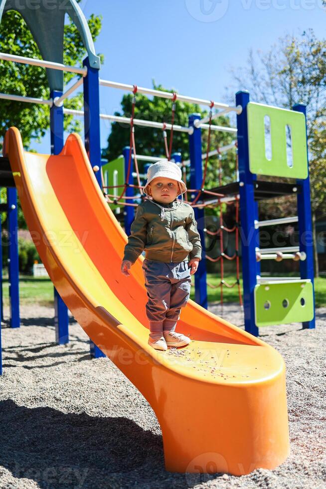 An adventurous, energetic toddler is joyfully playing on a bright orange slide situated at a vibrant playground on a sunny day, surrounded by the beauty of nature and opportunities for fun photo