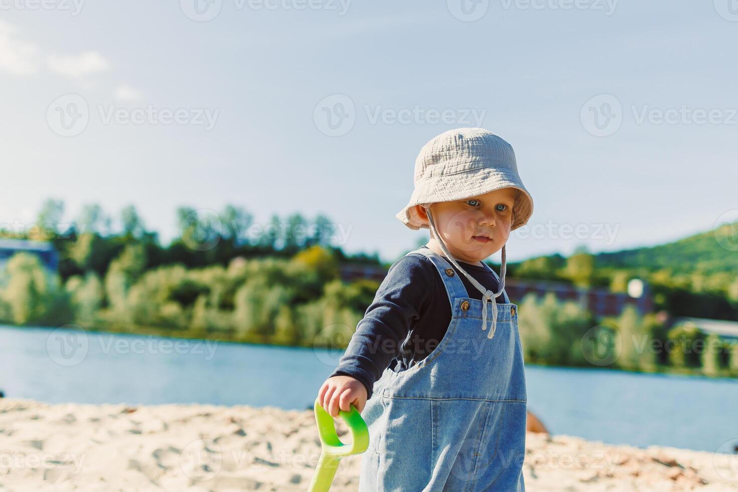 A Cherubic Toddler Enjoys a Beautiful Beach Day with a Brightly Colored Toy Shovel, Filled with Sunshine, Joy, Adventure, and Discovery with Family and Friends at the Tranquil Waterfront photo