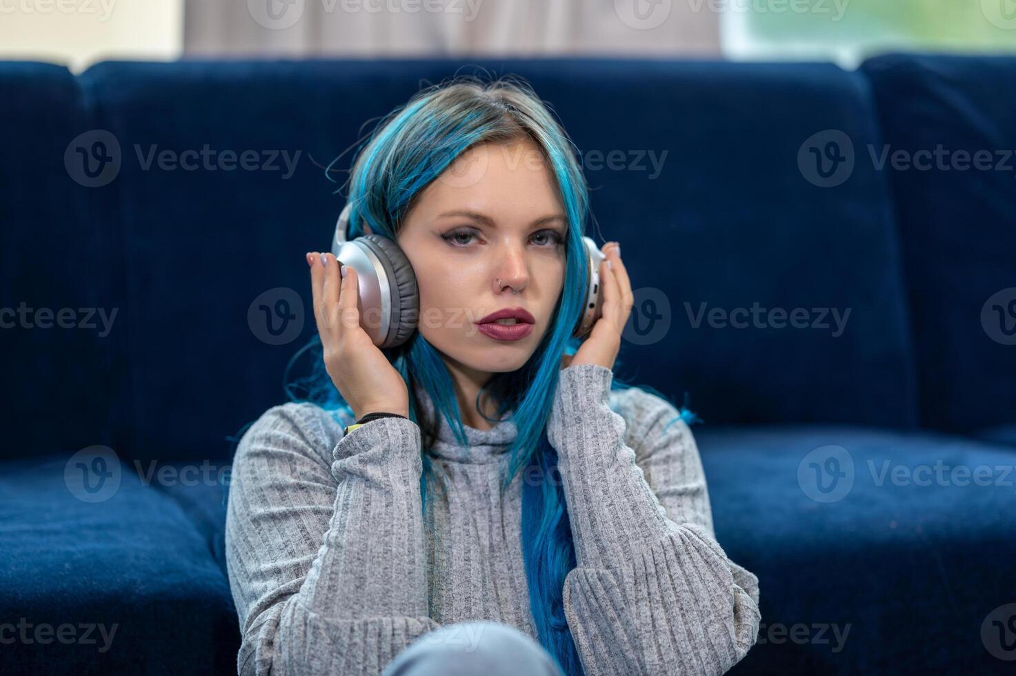Gorgeous portrait of a beautiful blue haired hipster listening to music on headphones while relaxing on a couch. Use for entertainment and technology. Look at the camera. photo