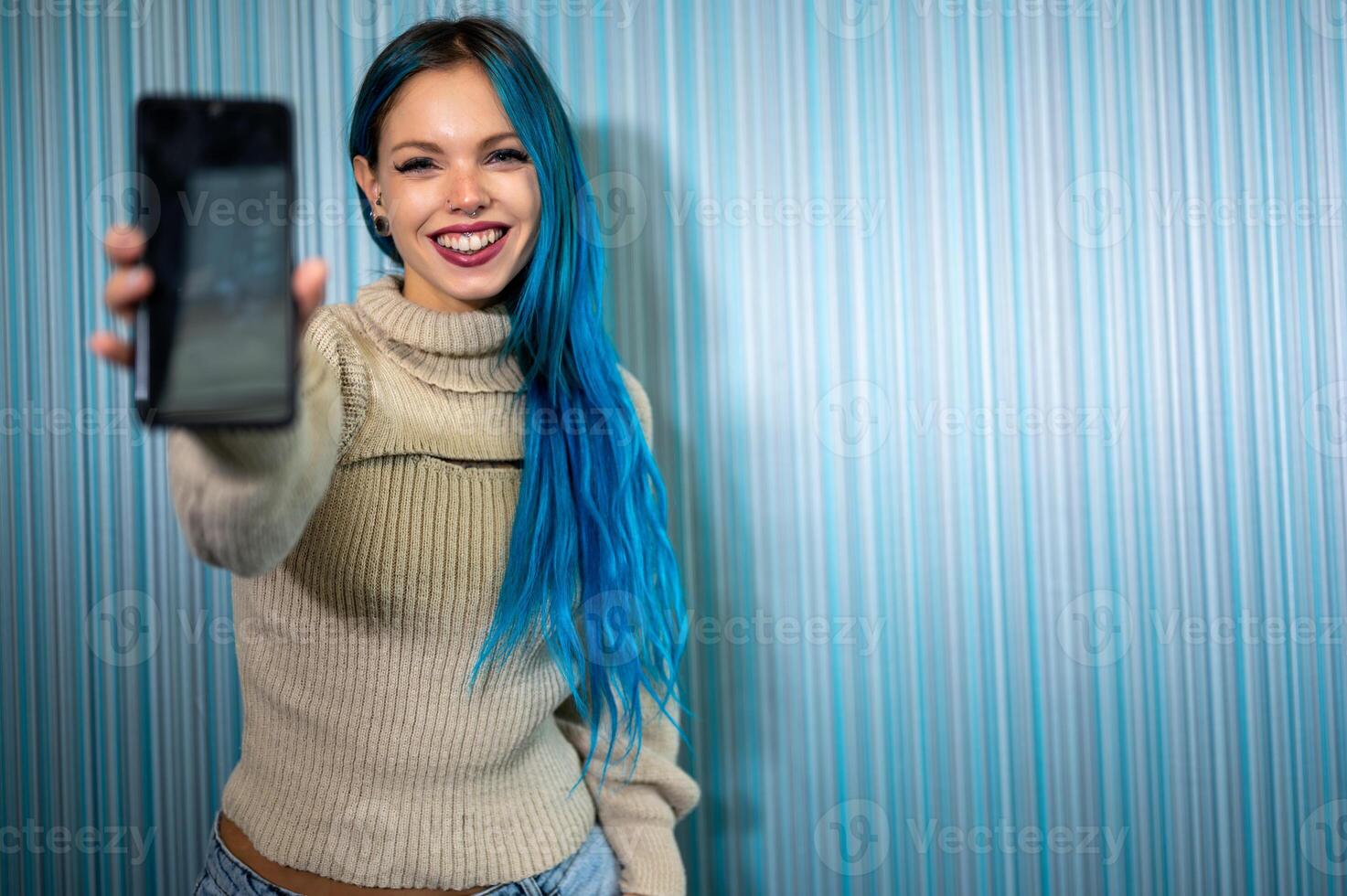 Portrait of a beautiful blue-haired hipster woman receiving a notification that causes her to have an extremely positive and surprised reaction. She extends her arm with her mobile phone towards us photo