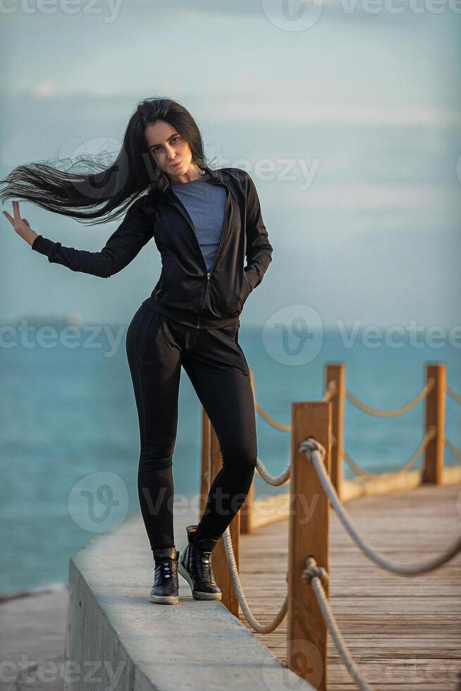 Woman posing confidently on a waterfront walkway during golden hour with flowing hair in a casual outfit photo