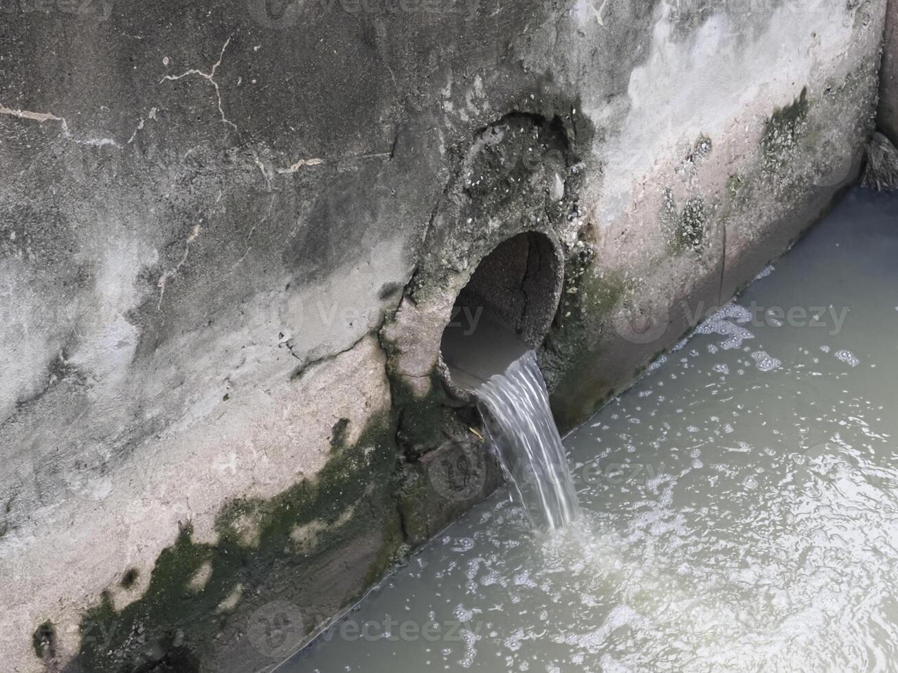 Small pipe embedded in weathered concrete wall releases water into murky water body, with moss and algae growth around pipe opening, creating natural, aged, and slightly neglected scene photo