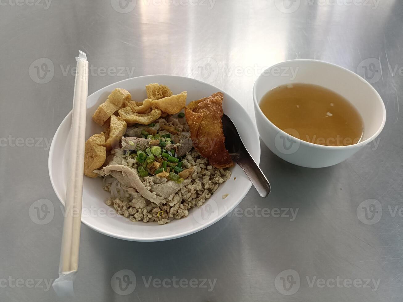 Delicious and comforting Asian food meal with noodle, meat, crispy skin, green garnish, in white bowl with hot savory broth soup on metal table, traditional street cuisine dish ready to eat photo