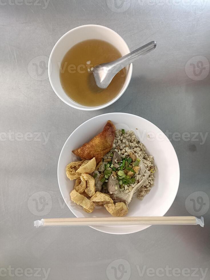 Bowl of clear broth with metal spoon plate with fried pork minced meat chopped green onions and crispy pork rinds along with pair of chopsticks all placed metallic surface creating simple photo