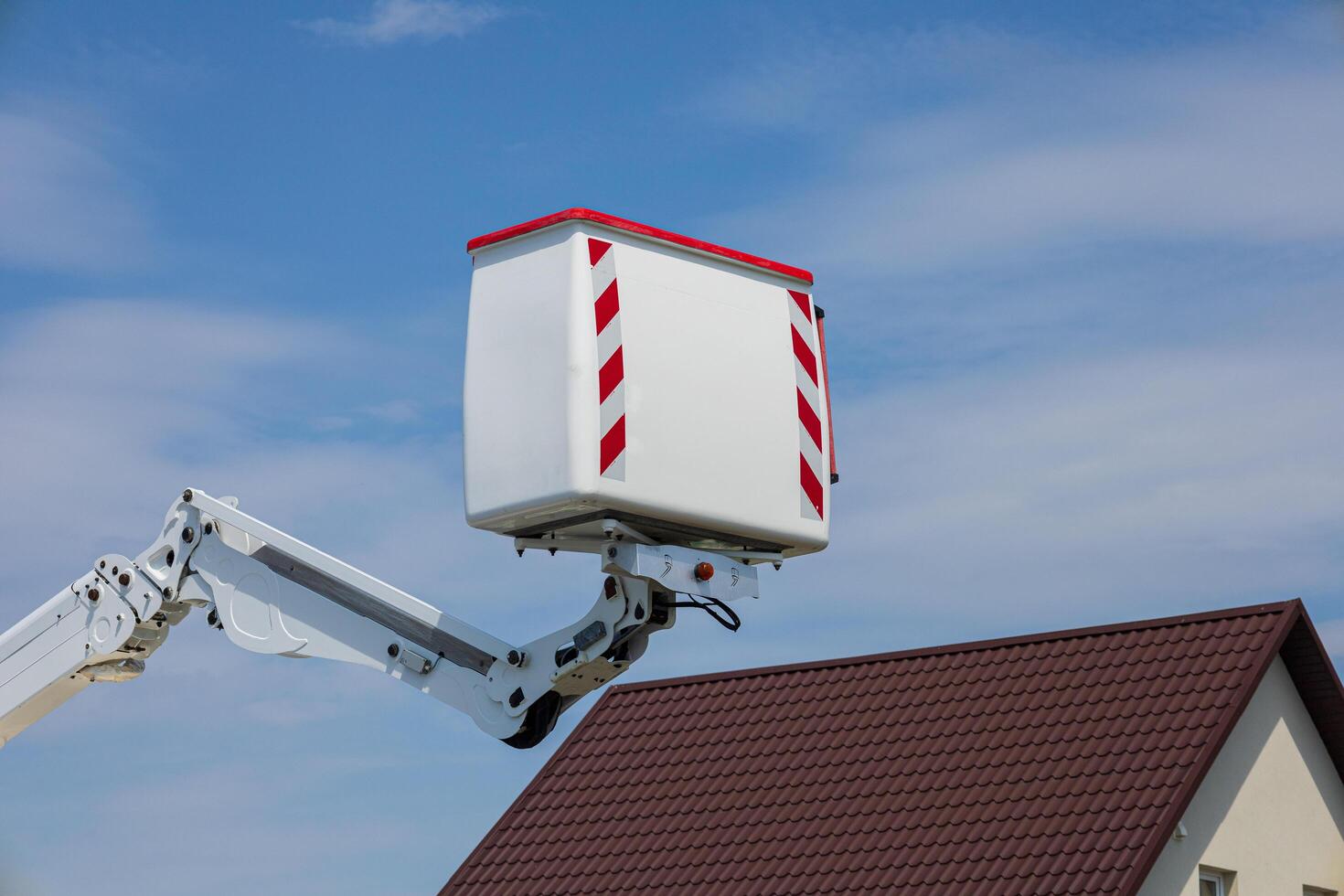 A worker operates an elevated platform to perform maintenance tasks on a residential roof in bright daylight with a picturesque sky photo