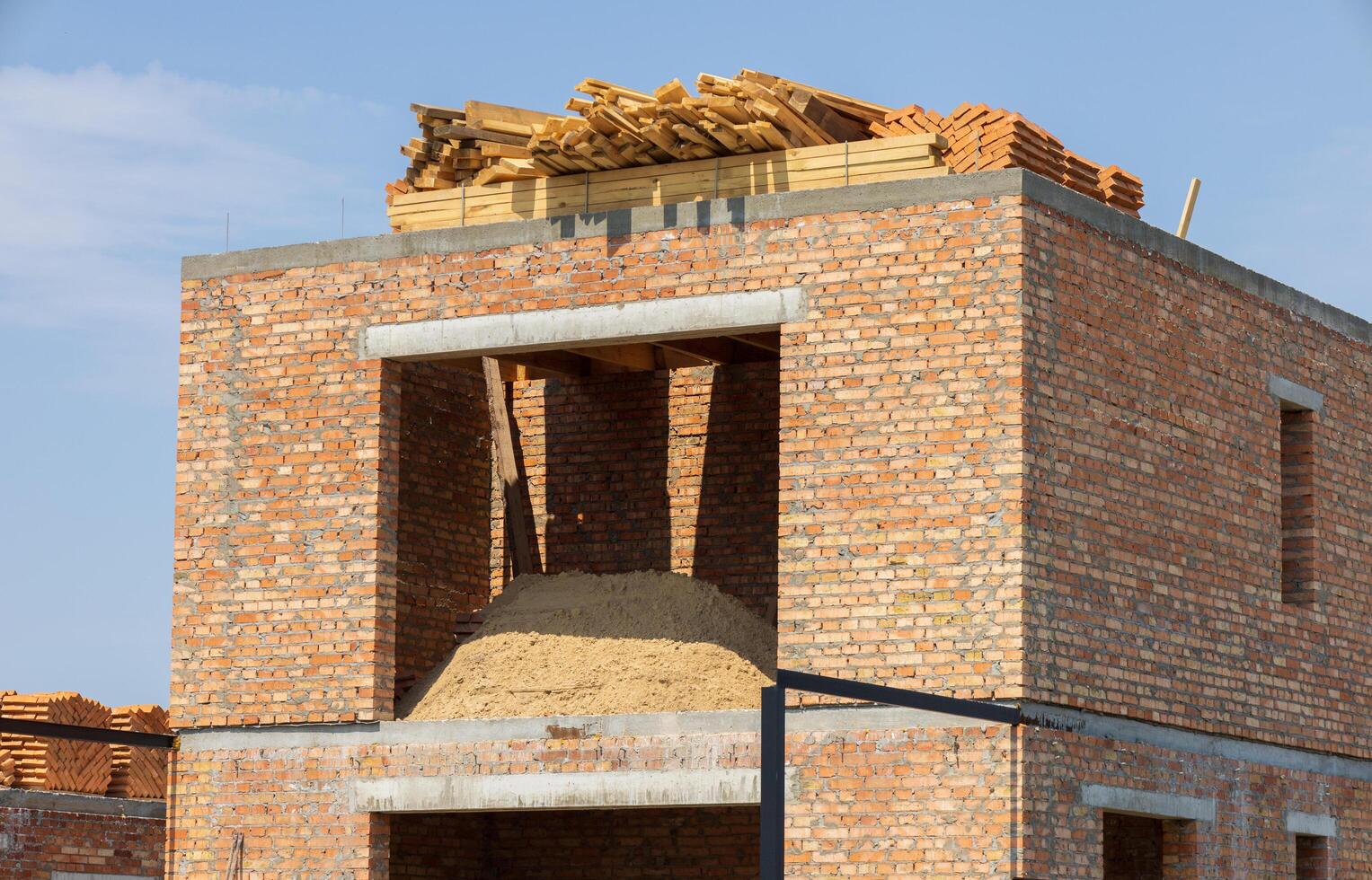Brick building is under construction, showcasing raw materials like sand and wooden planks on the rooftop against a sunny backdrop photo