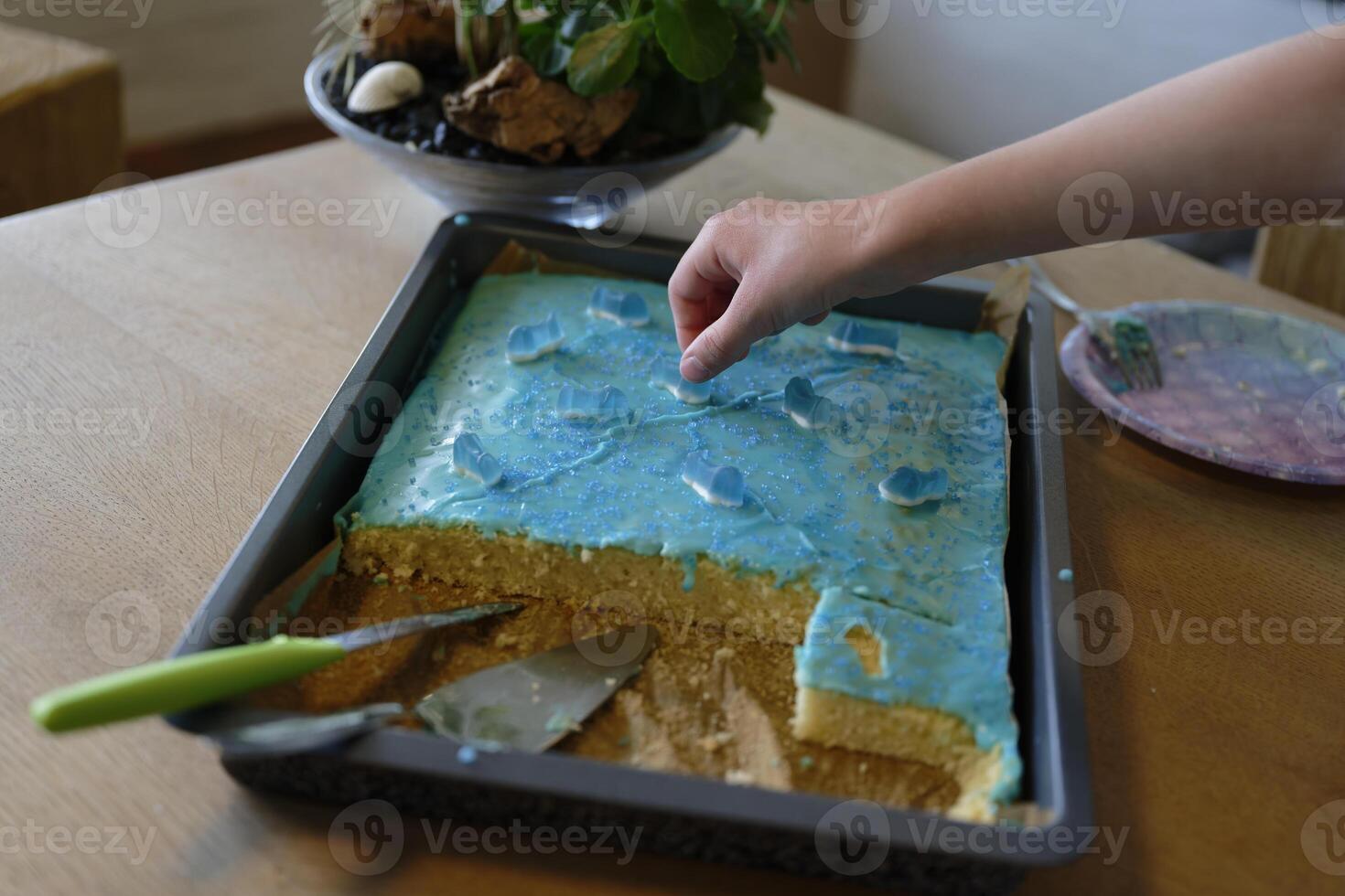 A childs hand reaches for a wine gum figurine on a blue sheet cake at a childrens birthday party photo