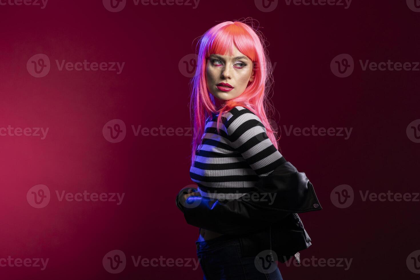 Female model with vibrant hair shows strong character in striped clothing while posing against isolated background. Pretty lady with pink wig presents clean confident stance inside fashion studio. photo