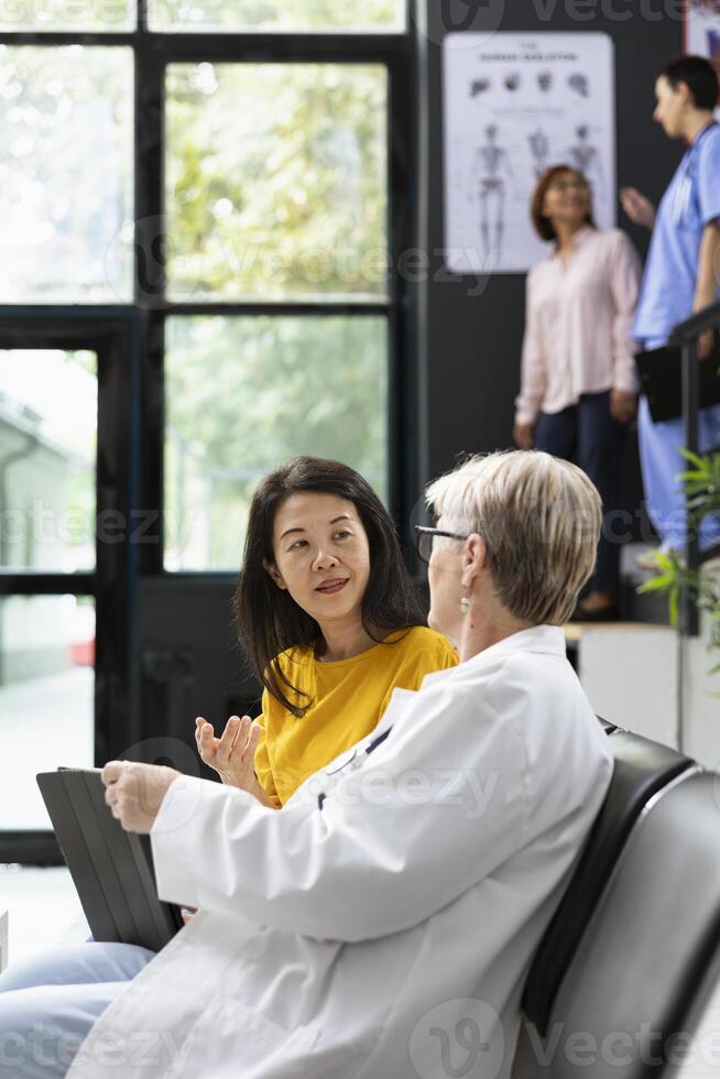 In a modern lobby, physician speaks with asian woman about upcoming medical tests. Experienced medic having a conversation for health prevention, test results evaluation and wellness. photo