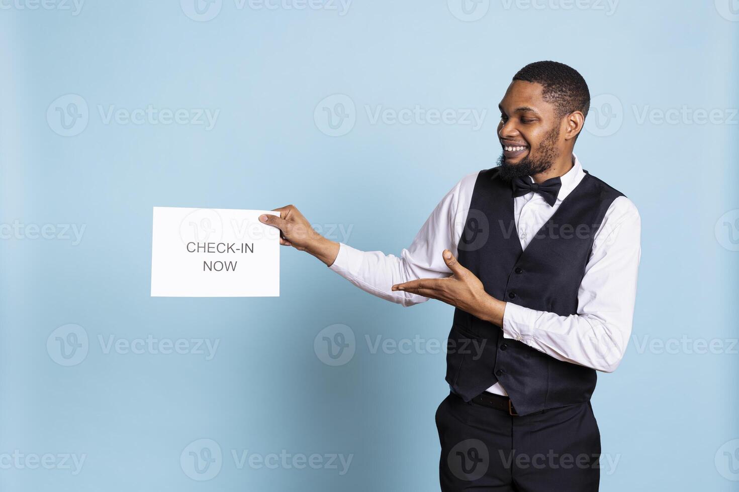 Professional bellboy showing a paper sign for guests to go to room check in, presenting an indicator to get the attention of clients. Hotel concierge in a tuxedo standing against blue background. photo