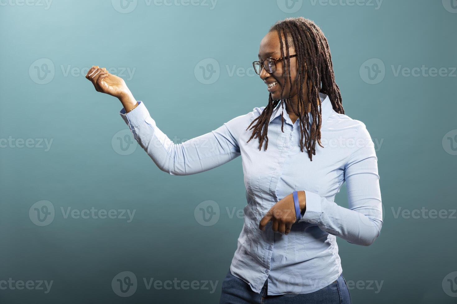 Smiling black woman with glasses having a good time in blue background studio. African american female individual standing and playing an imaginary guitar, feeling happy and optimistic for new day. photo