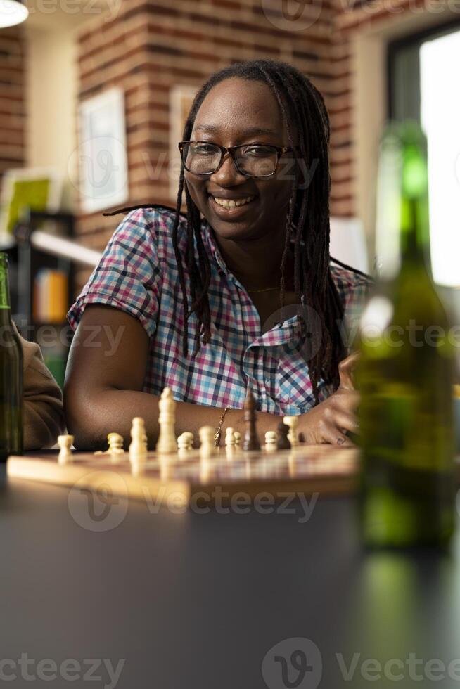 Selective focus on african american woman watching an ongoing friendly chess match on table. Smiling black lady with glasses sitting and enjoying board game session in brick wall room. photo