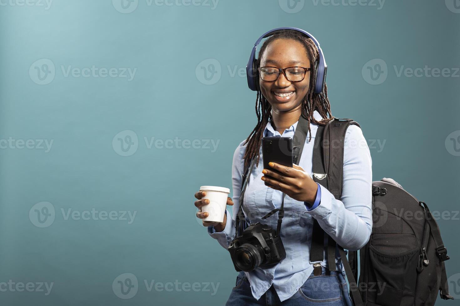 Joyful black woman with headphones watching funny videos on smartphone as she stands with coffee cup in hand and dslr camera around her neck. Female photographer using mobile phone with a smile. photo