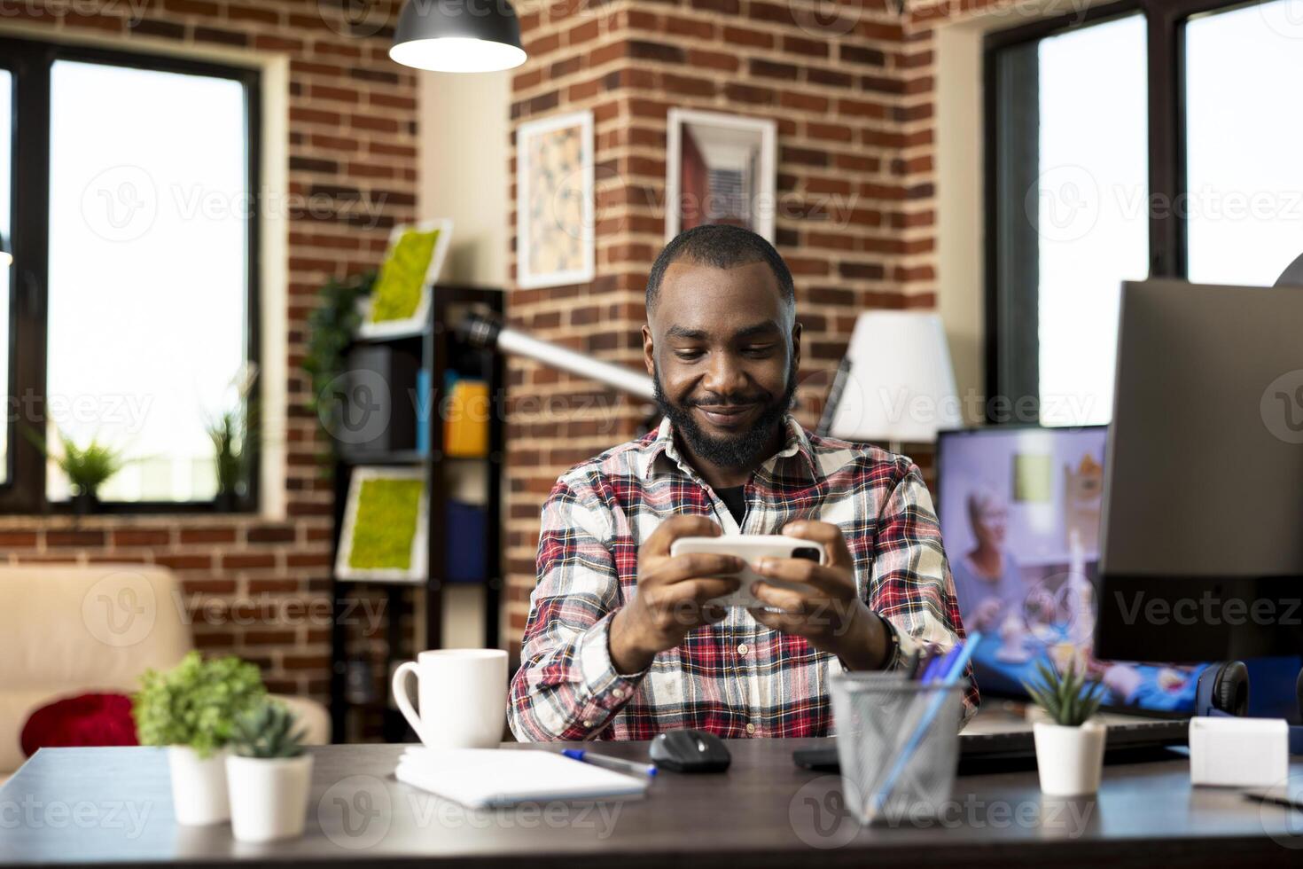 Smiling african american man sitting at desk, enjoying mobile game on smartphone during break time. Happy remote worker with digital device in hand, relaxing in modern home office with joy. photo
