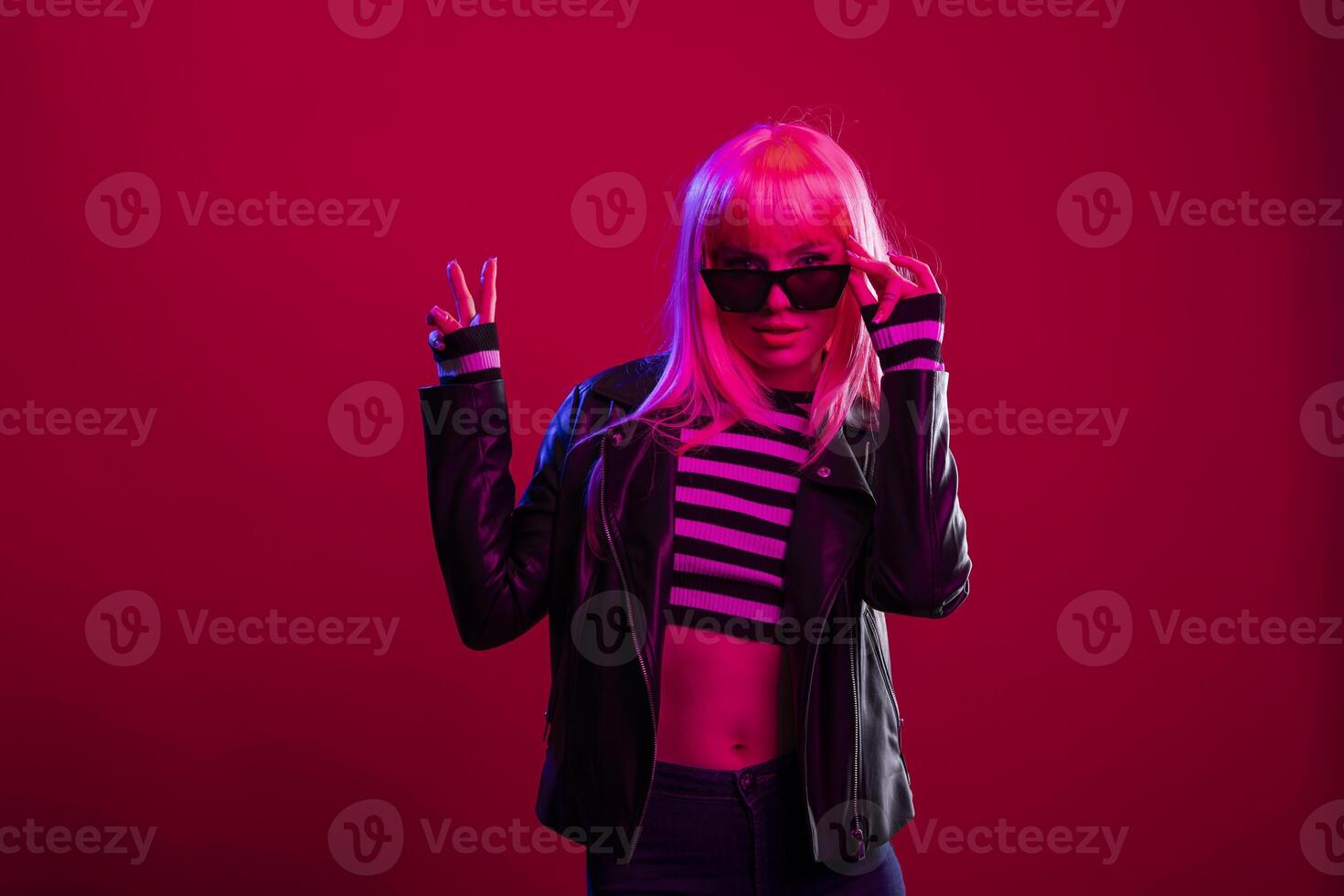 Confident glamorous woman in leather jacket and sunglasses modelling with carefree punk energy in studio. Funky female model touching her black shades and posing with peace sign hand gesture. photo