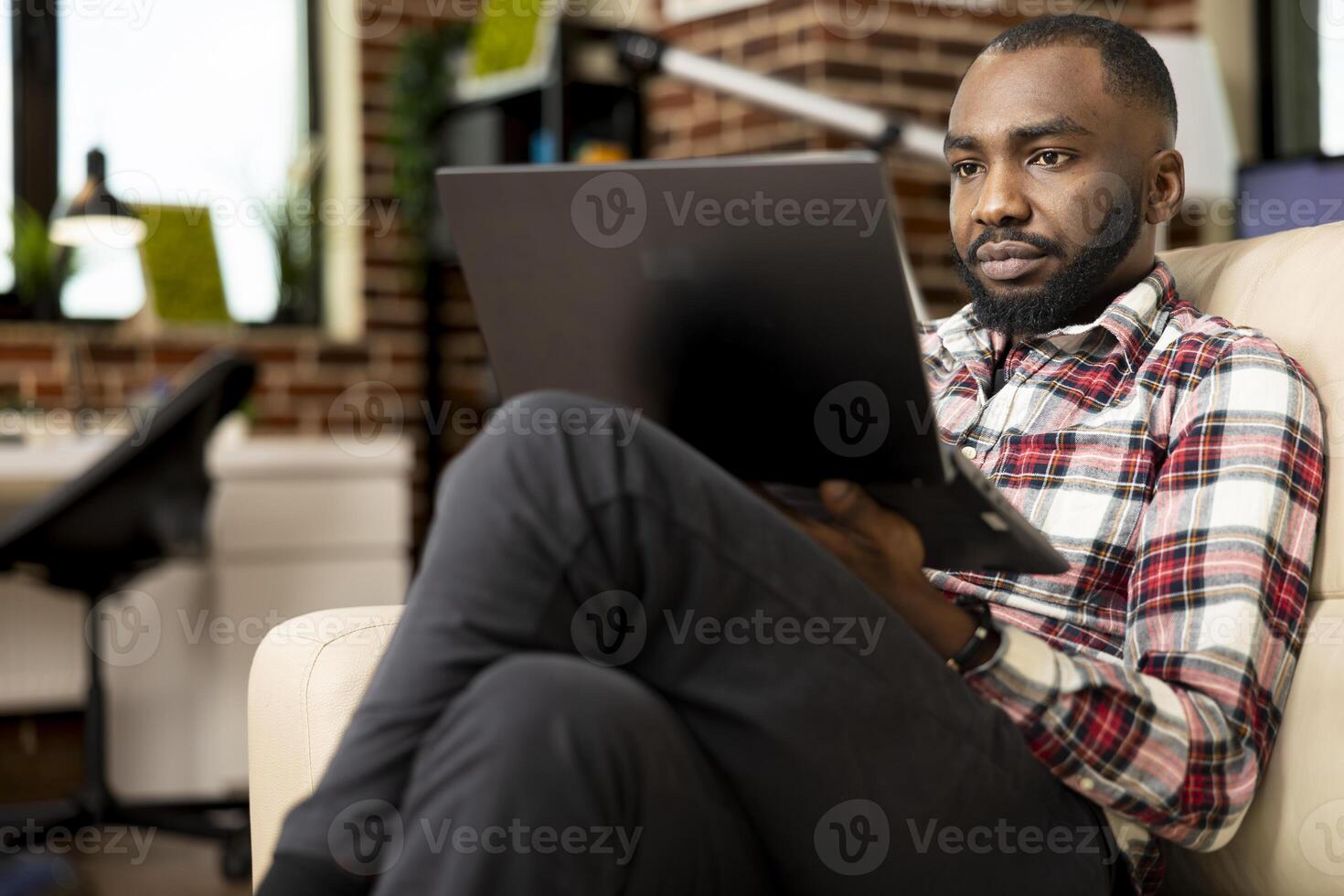 African american businessman seated with legs crossed, working from home, preparing startup project reports. Focused freelancer uses laptop on couch, updating resume and browsing job opportunities. photo