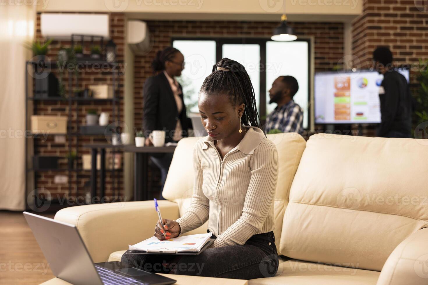 Black female employee reads documents from clipboard, combining digital tools and paperwork to prepare a business plan. African american businesswoman seated on office sofa, reviewing printed reports. photo