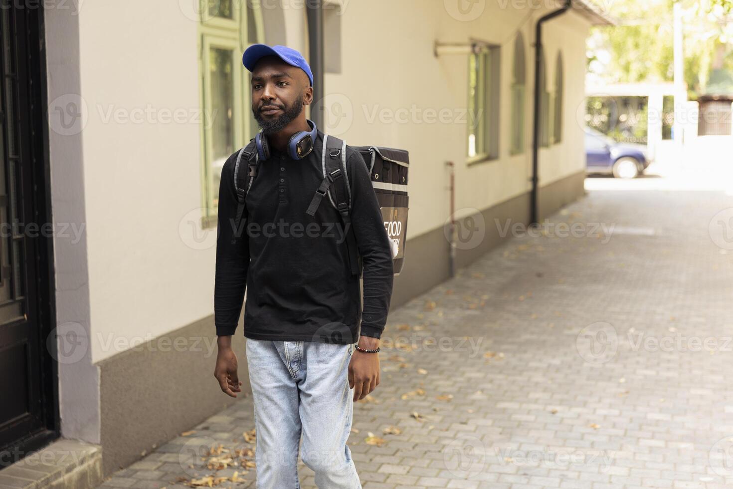 Black male courier approaches the front door to drop off a hot meal order, ringing the intercom to deliver the food. Young adult employee ensuring an express delivery takeaway service. photo