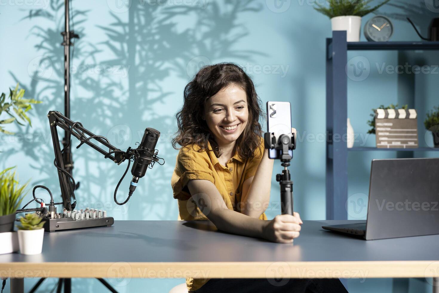 Happy influencer in home studio greeting audience, recording live broadcast using smartphone on selfie stick. Woman filming q and a session with mobile phone, reading questions from fans photo