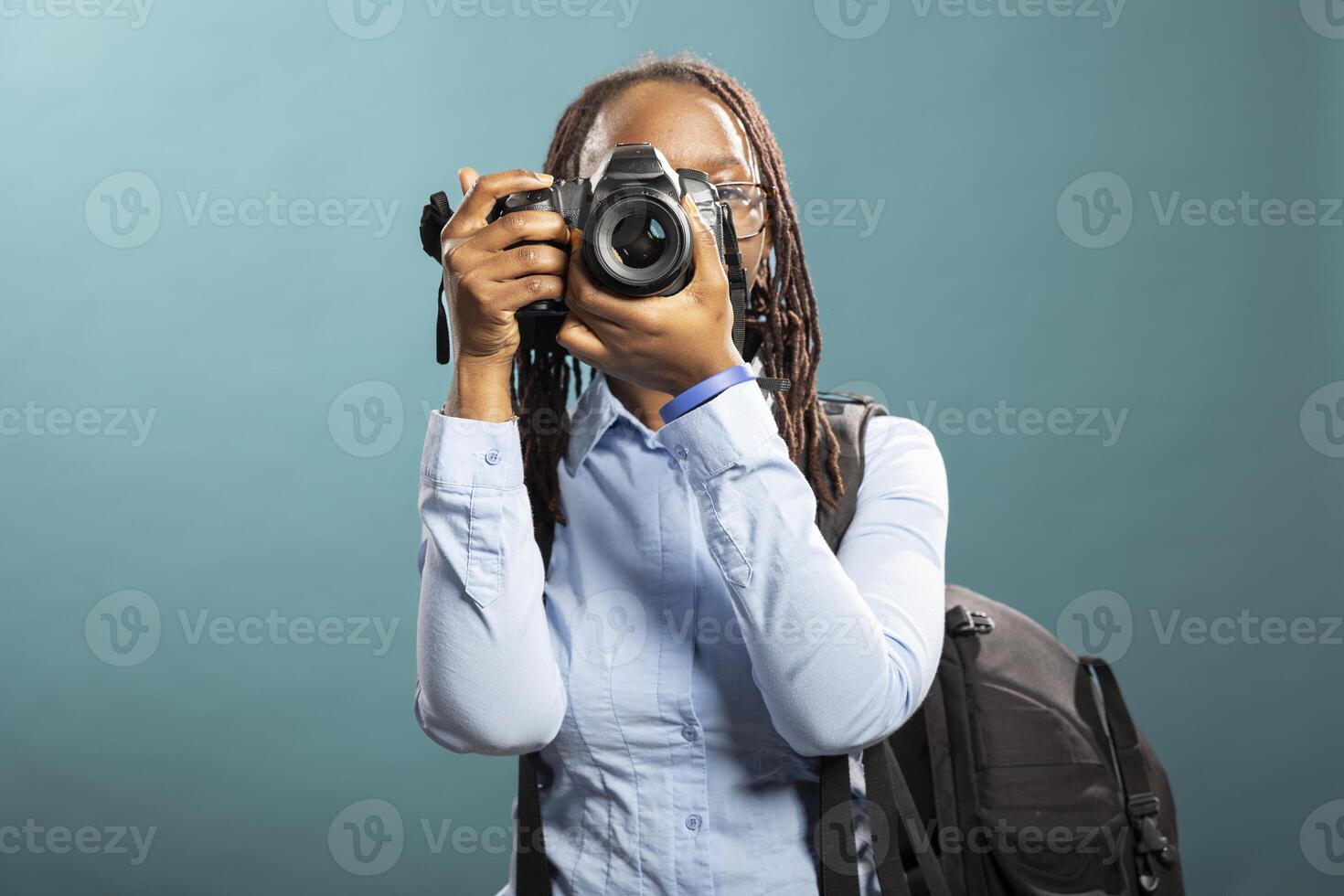 African American woman wearing backpack, prepares to shoot, holding DSLR near her face in confident, instructional pose. Black female photographer shows how to use camera, holding it close to eye. photo
