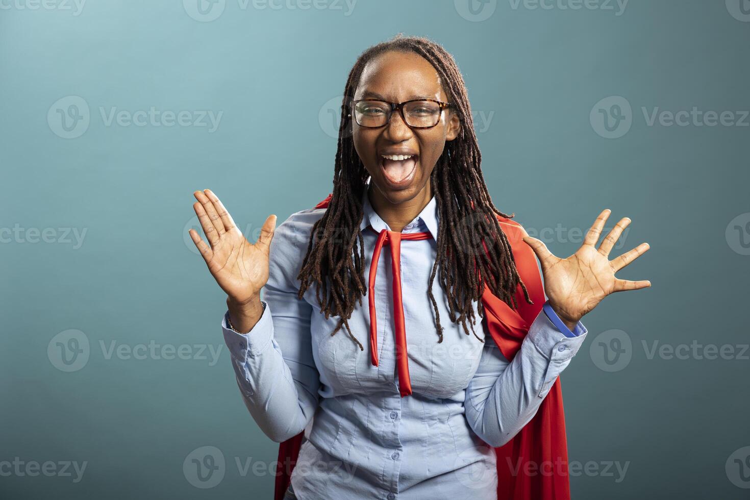 Confident black woman wearing a red cape and blue shirt, expressing authentic excitement in modern studio. African american heroine with her hands and mouth wide open, shouting with joy. photo
