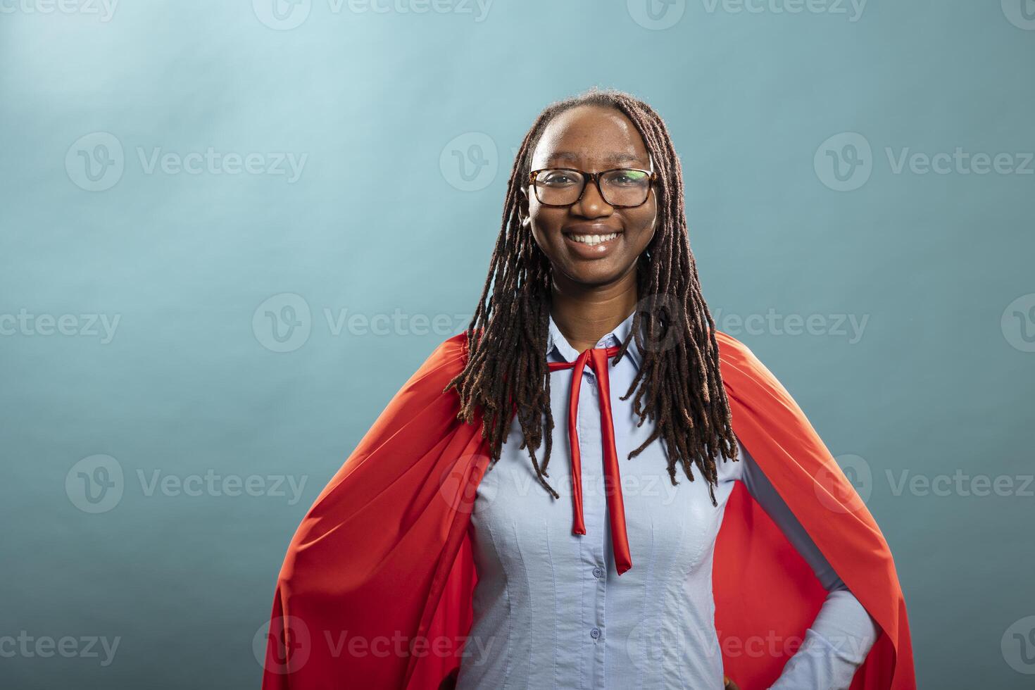 African american woman with red cape posing in blue background studio and confidently smiling at camera. Portrait of black female superhero standing strong, feeling empowered. photo